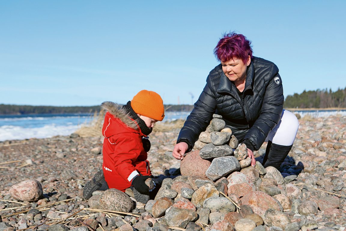 Isoäiti ja lapsenlasi kasaavat rannalla kiviä keoksi. Sininen taivas. 