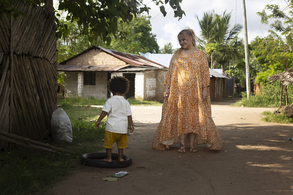 Kuva Viola Wanniuksesta kävelemässä hiekkatiellä poikansa kanssa. Taustalla on trooppisia puita ja yksinkertainen talo.