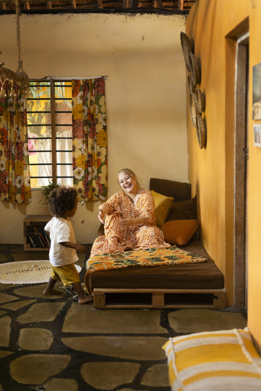 Viola istuu matalalla sohvalla ja nauraa poikansa juoksentelulle. Taustalla on keltainen huone.