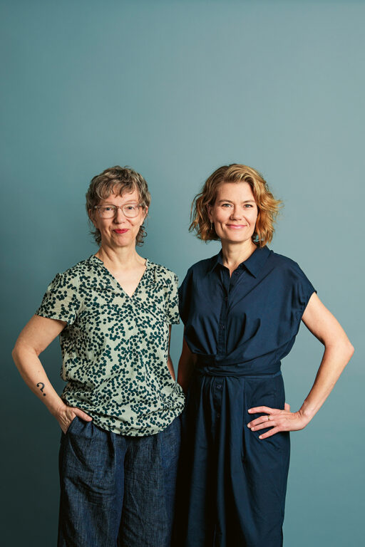 Pauliina Pulkkinen ja Leena Virtanen seisovat sinistä taustaa vasten studiokuvassa.