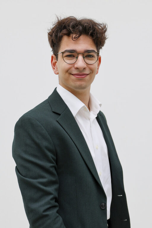 Potretti Andreas Dimitriousta, joka seisoo puku päällä valkoista taustaa vasten. Hänellä on ruskeat hiukset ja silmälasit.