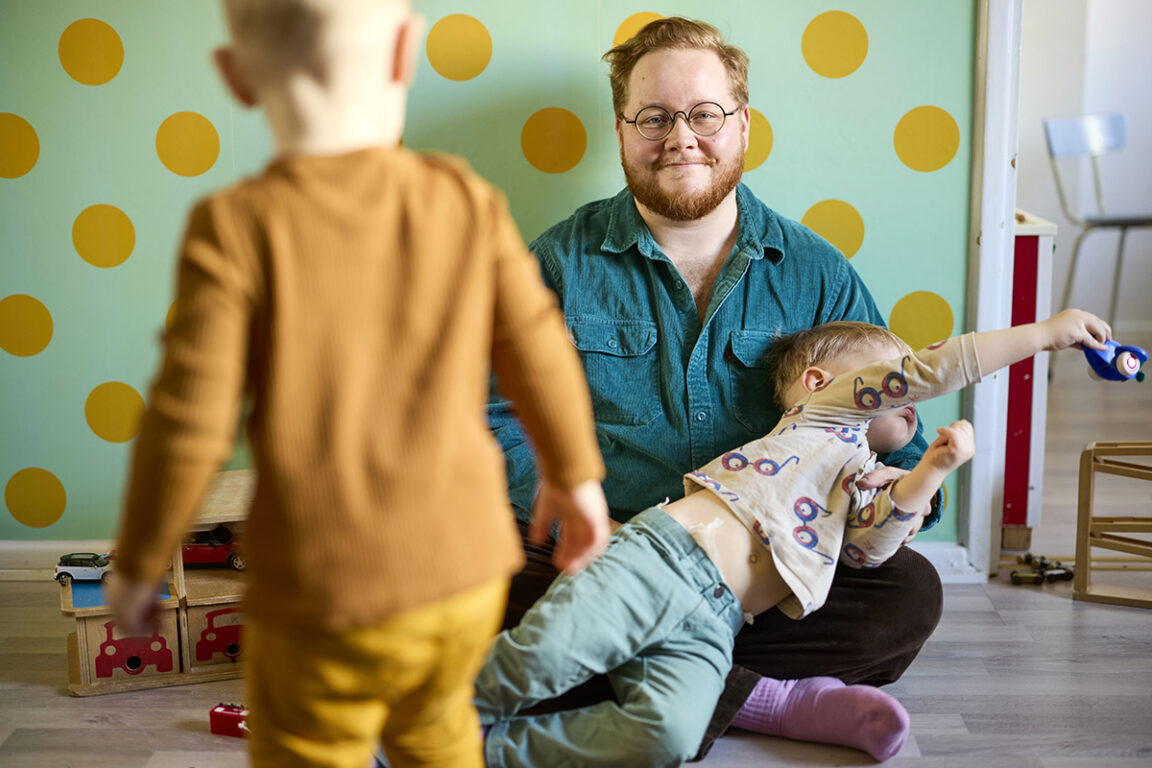 Nooa Sammalkäpy istuu lattialla ja pitää sylissään temppuilevaa taaperoa. Etualalla on selin toinen taapero. Taustalla näkyy värikäs koti.