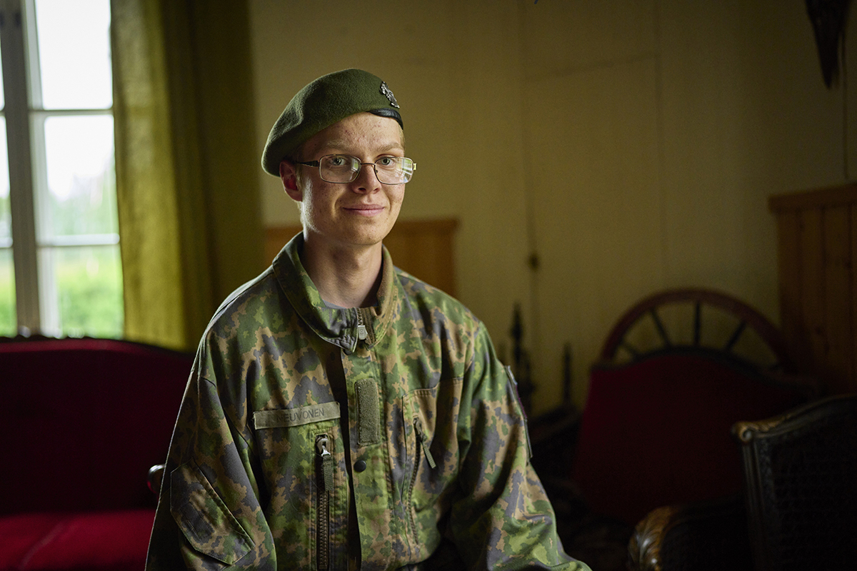 Nuori mies istuu kotonaan armeijan varusteet päällä ja hymyilee kameralle.