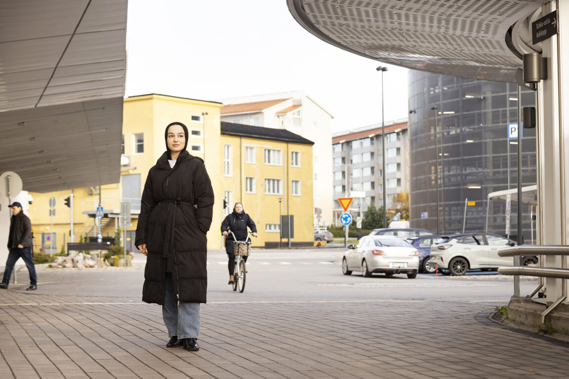 Fatib seisoo rakennusten kattojen välissä kadulla. Taustalla näkyy ihmisiä ja autoja.