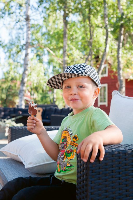 Lippispäinen pieni poika istuu hymyillen tuolilla ulkona ja syö jääteloä.