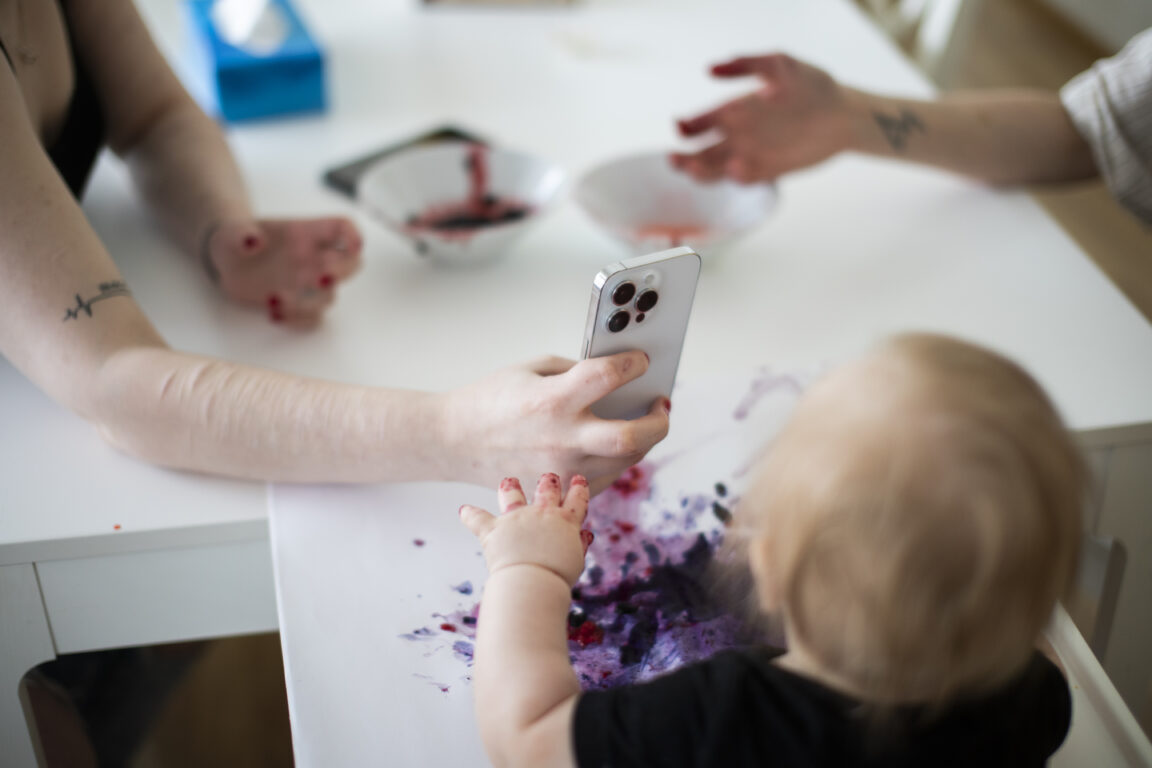 Kuvassa nuoren ihmisen käsi pitelee puhelinta ja ottaa kuvaa vauvasta, joka piirtää mustikkasoseella.