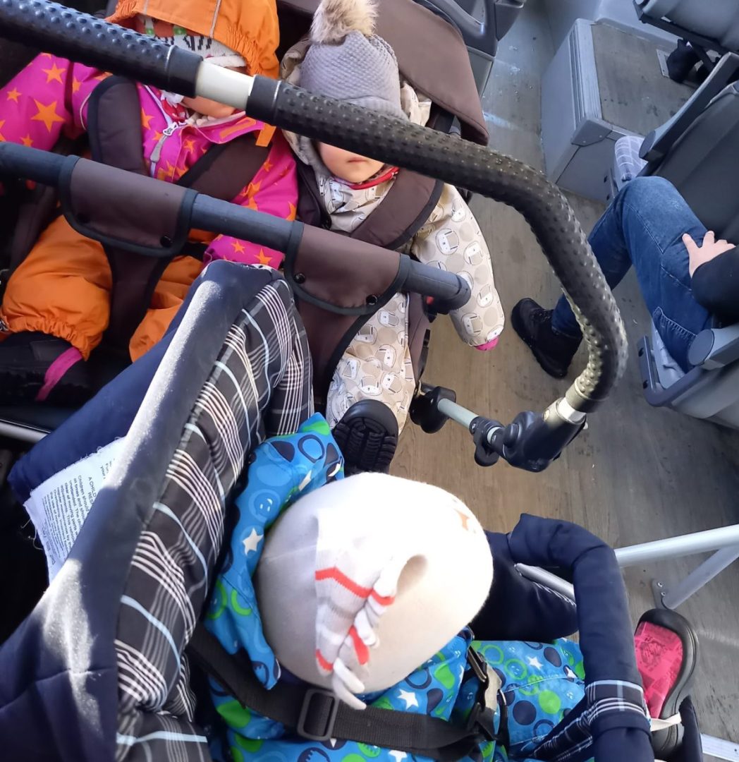 Kolme lasta kaksissa eri kärryissä bussissa. Kasvot eivät näy.