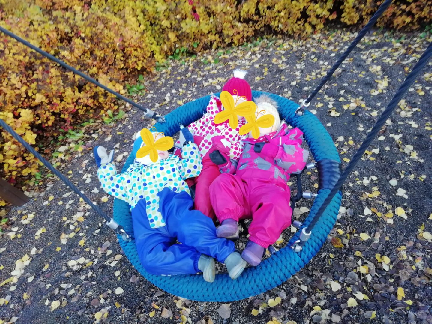 Kolme lasta spiderkeinussa. Kasvot peitetty emojein.
