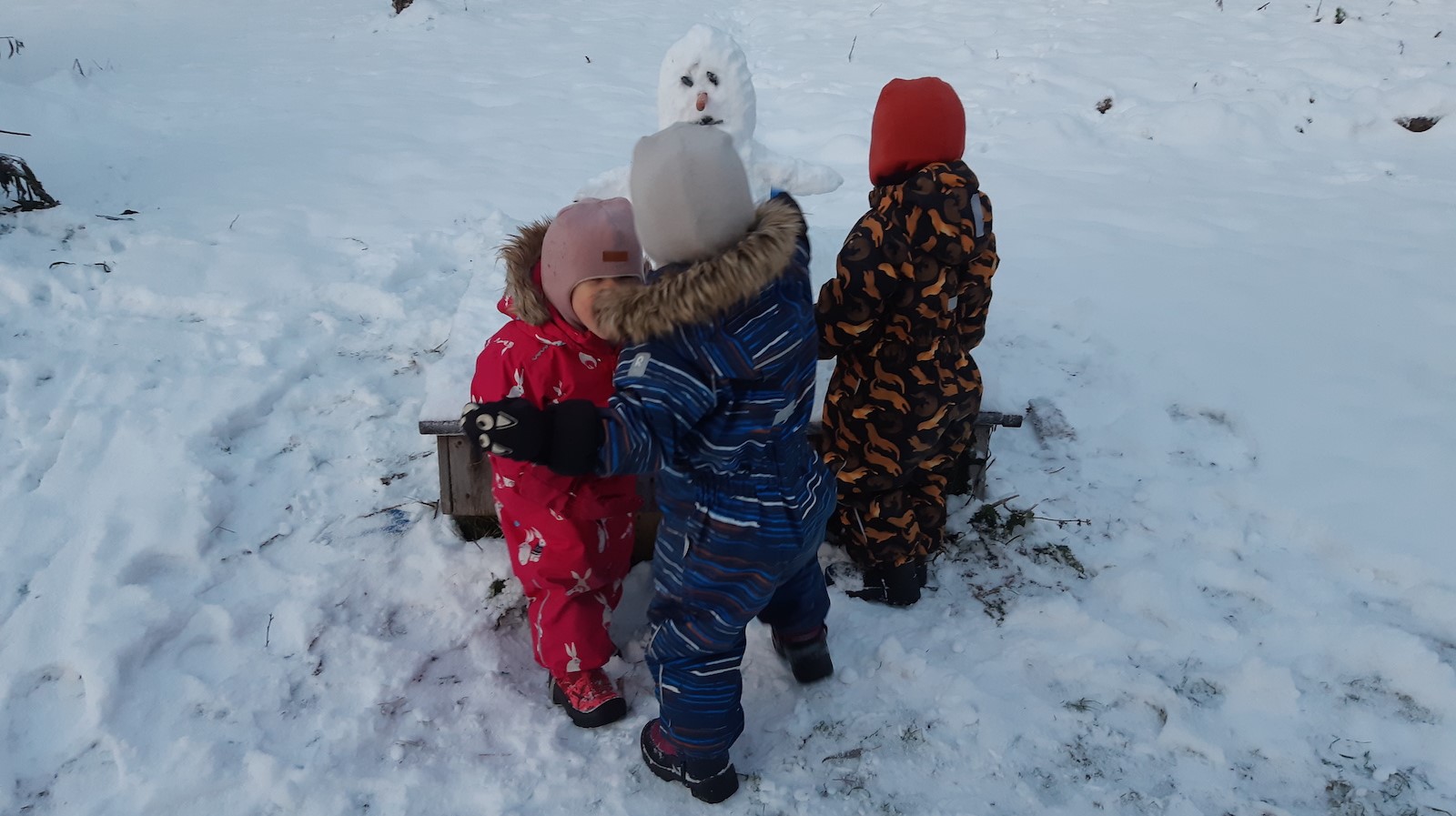 Kuvassa on kolme taaperoa ulkona leikkimässä lumiukon edessä.