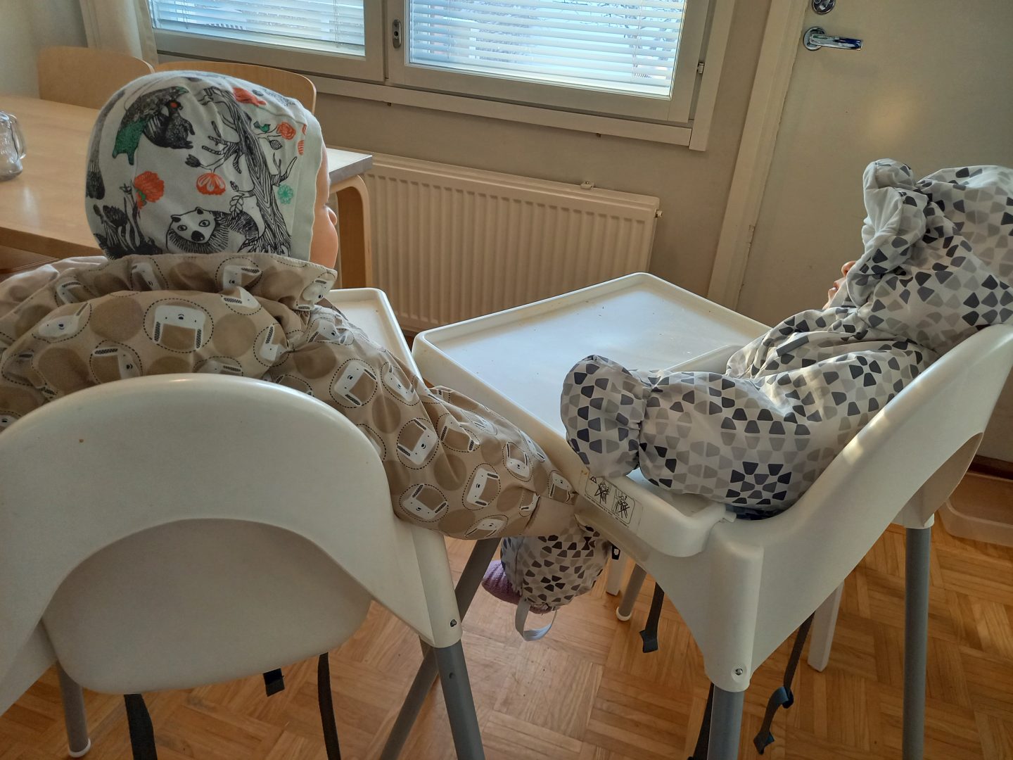 Kaksi lasta toppapuvuissa syöttötuoleissa istumassa, takaapäin.