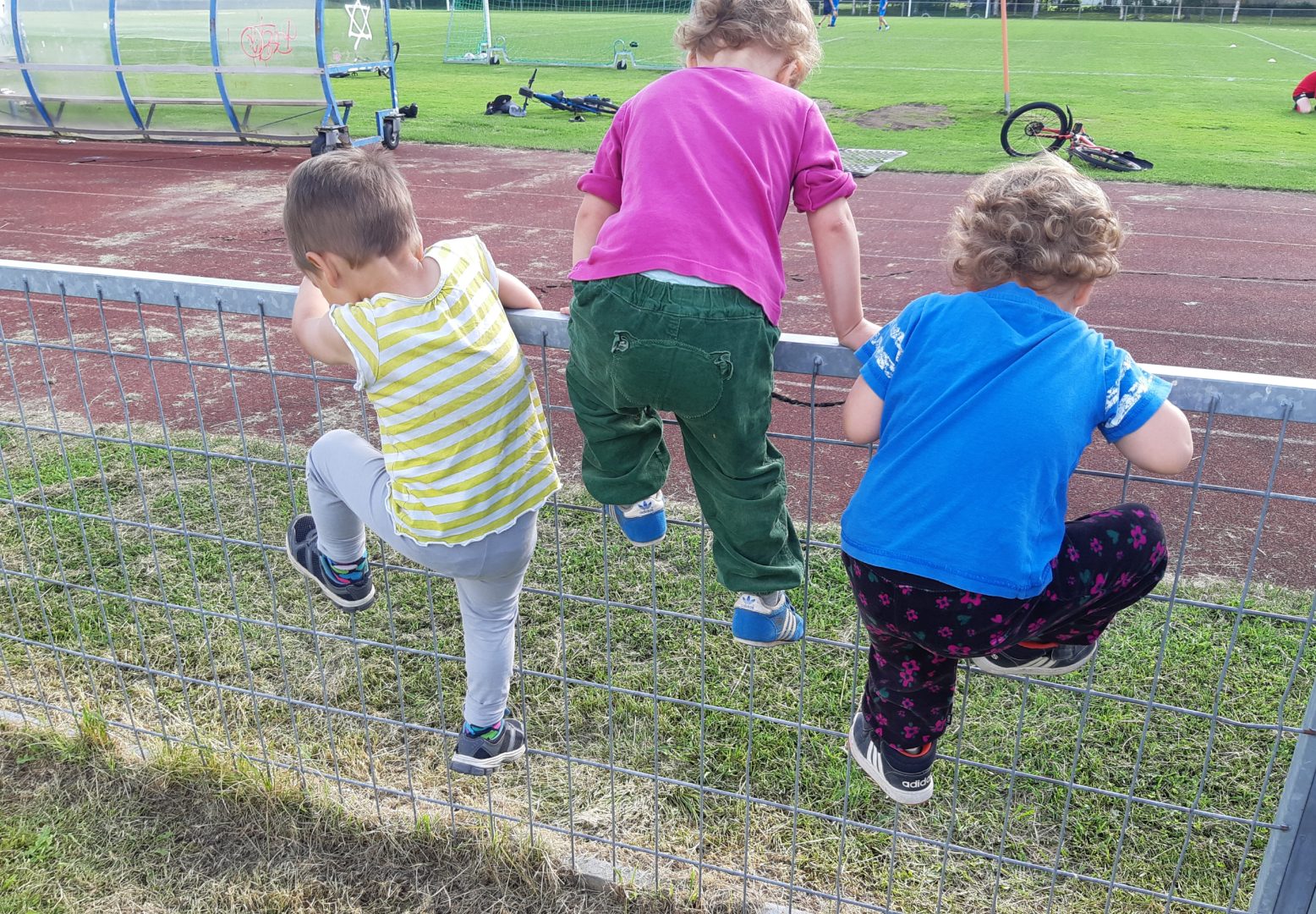 Kolme lasta kiipeilemässä aitaa ylös jalkapallokentän reunalla.