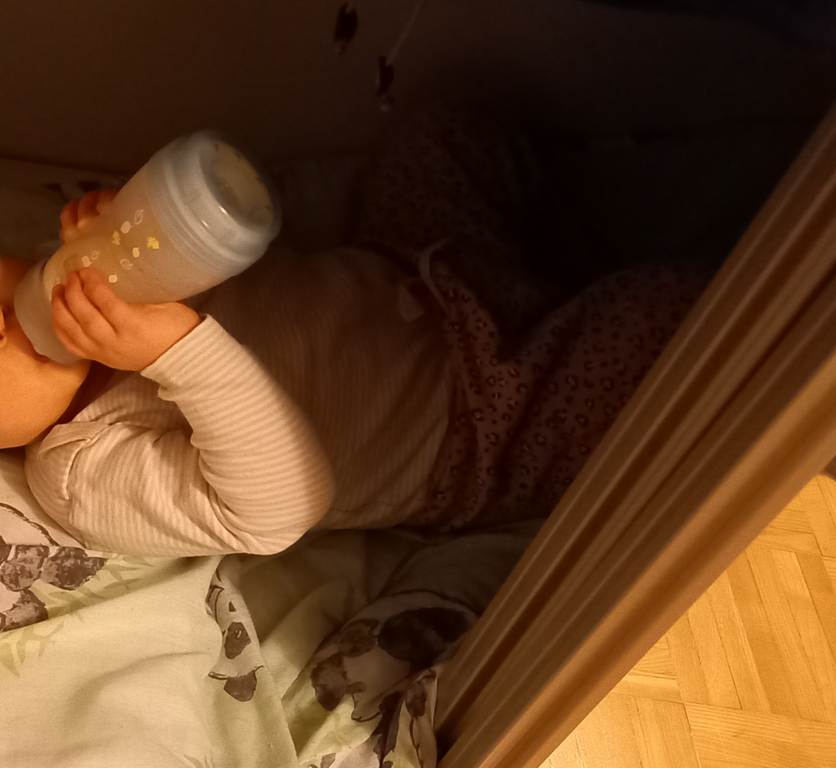 Pikkulapsi makoillee selällään eteisen komerossa tuttipullo suussa. Vain kasvojen alaosa ja osa vartaloa näkyy.