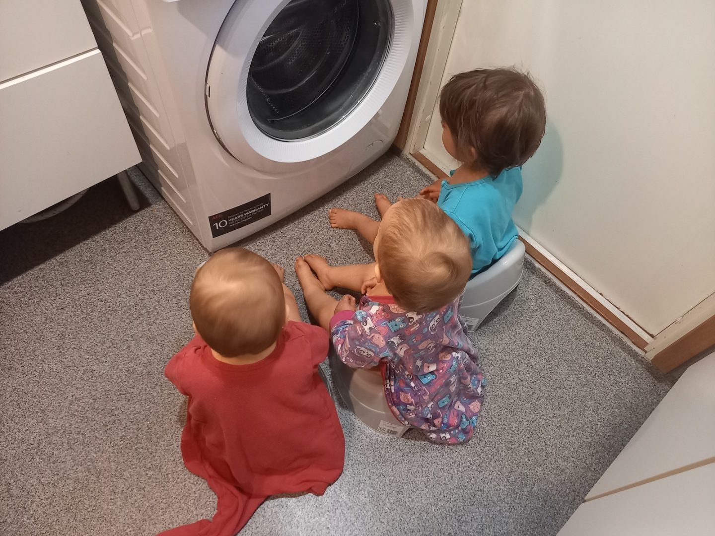 Kolme lasta takaapäin kuvattuna. Istuvat potalla yökkärit päällä pyykinpesukoneen edessä.