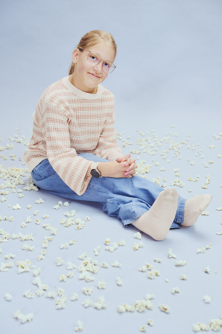Milja istuu studion lattialla sinistä taustaa vasten. Lattialla on popcorneja.