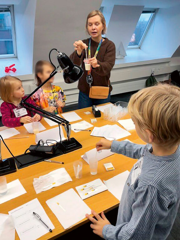 Lapsijoukko on kerääntynyt pöydän ääreen, missä nainen opastaa heitä pipetin kanssa.