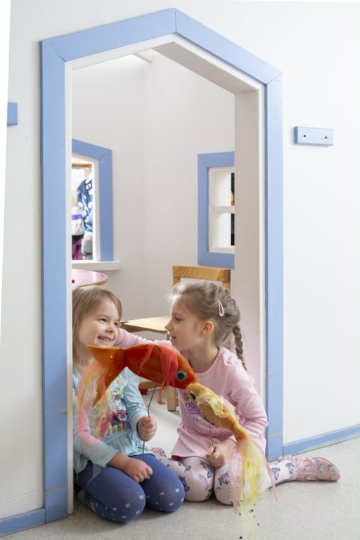 Kaksi iloista lasta istuu oviaukossa kahden ison leikkikalan kanssa.
