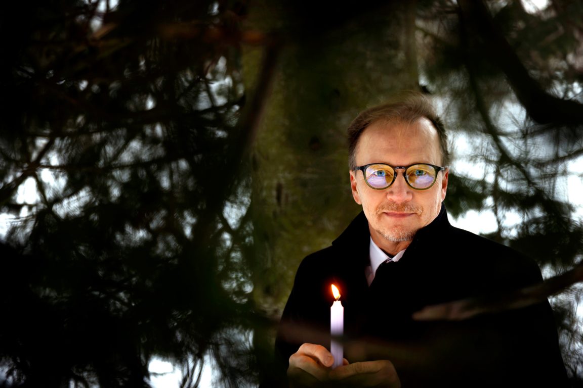 Jarmo Kokkonen seisoo suuren männyn alla kynttilä kädessään.