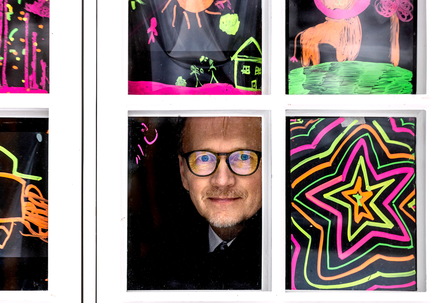 Jarmo Kokkonen kurkistaa ulos ikkunasta kameraa kohti värikkäiden piirustusten ympäröimänä.