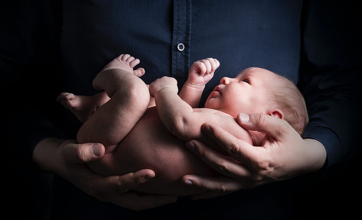 Mies pitää tummaa taustaa vasten alastonta pientä vauvaa sylissään.