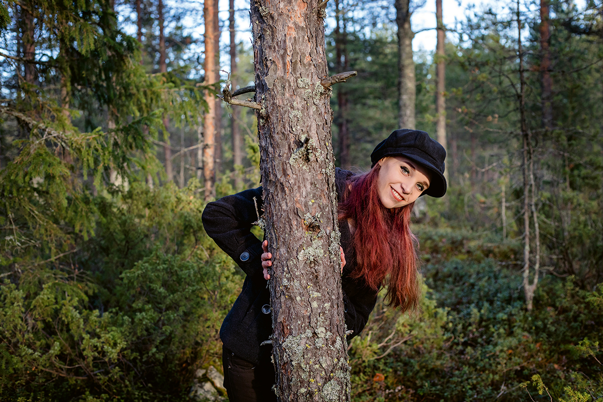 Iloinen, punahiuksinen tyttä kurkkaa metsässä puun takaa.