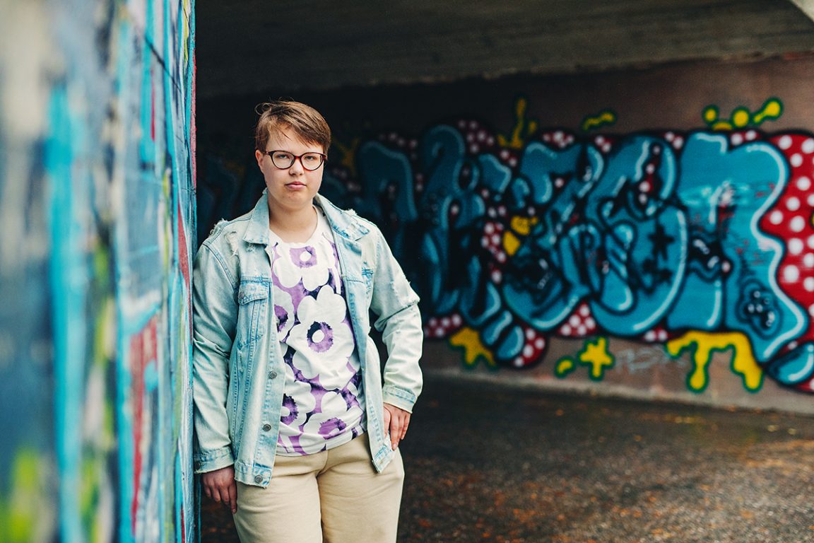Lyhyttukkainen, silmälasipäinen Riina Aho nojailee graffitilla peitettyyn seinään. 