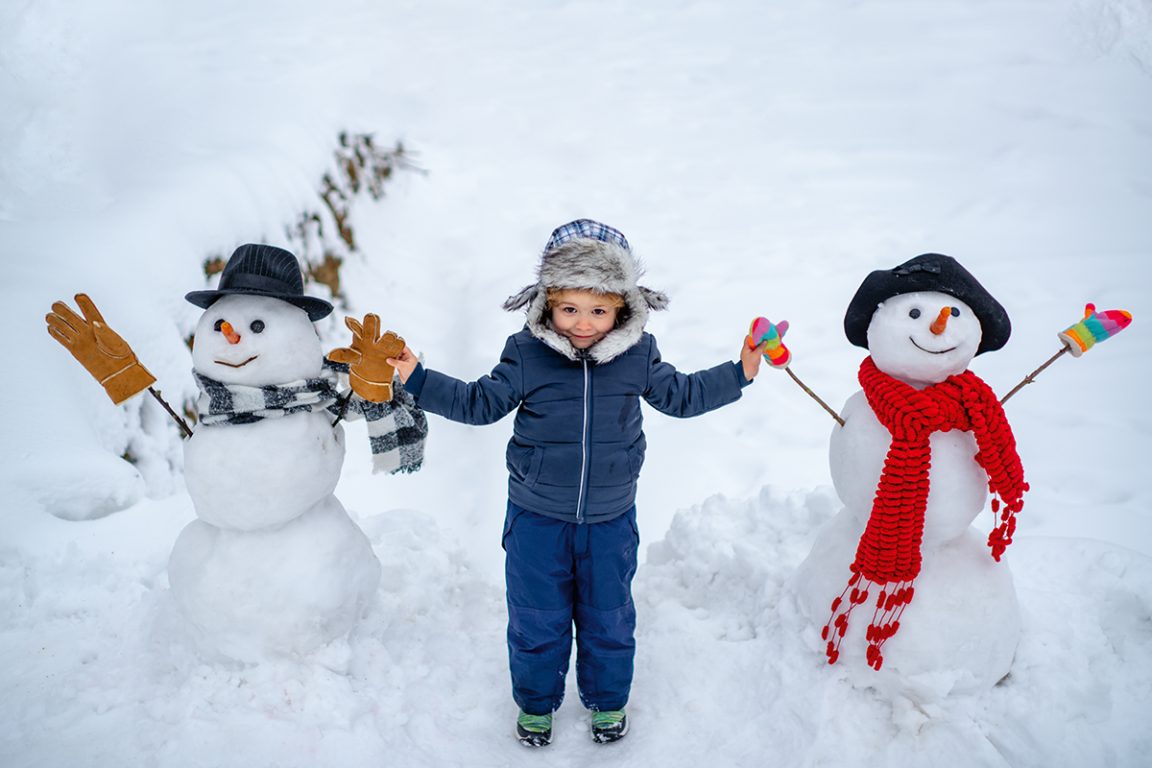 Lapsi seisoo iloisena lumessa kahden lumiukon välissä ja pitää niitä käsistä.