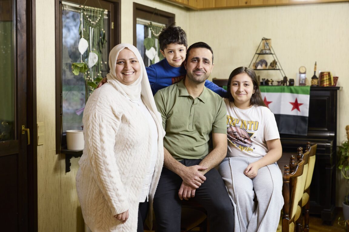 Nelihenkinen perhe katsoo kameraan kodissaan ja hymyilee.