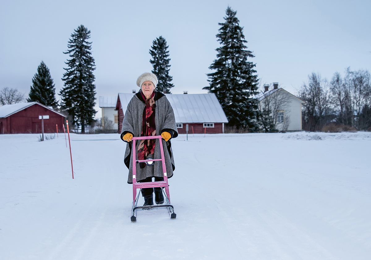Tuula Paltakari-Piekkola seisoo potkukelkan ohjaksissa talvisen maalaistalon pihalla.