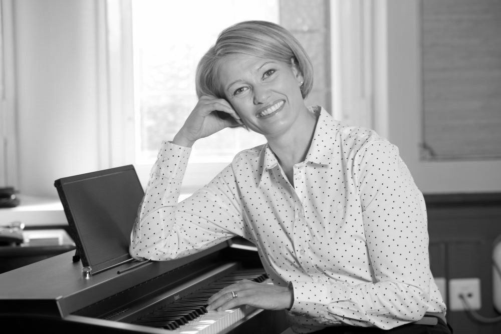 Ulla Siimes nojaa pianoon ja katsoo kameraa kohti hymyillen