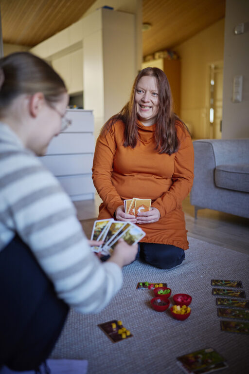 Kaksi naista, nuorempi ja vanhempi, pelaavat korttia omakotitalon olohuoneen lattialla.