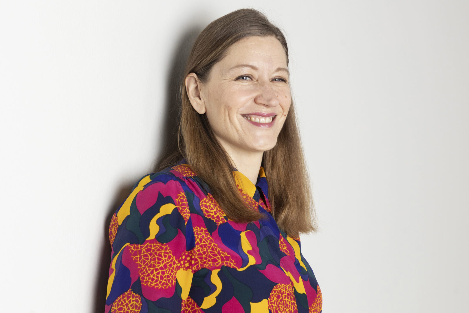 Merja Mähkä seisoo vaaleaa taustaa vasten ja hymyilee.