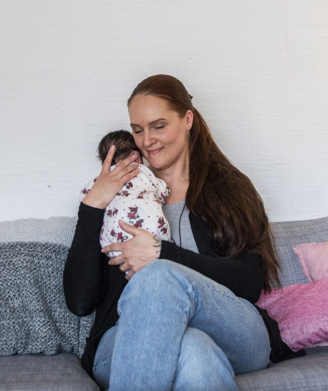 Ohjaaja pitää sylissään pientä vauvaa ja hymyilee silmät suljettuina.