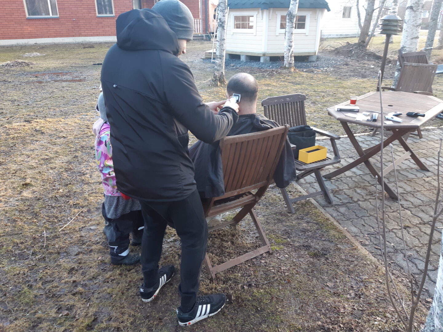 Mies leikkaa pihalla toisen hiuksia, lapset katsovat vierestä.