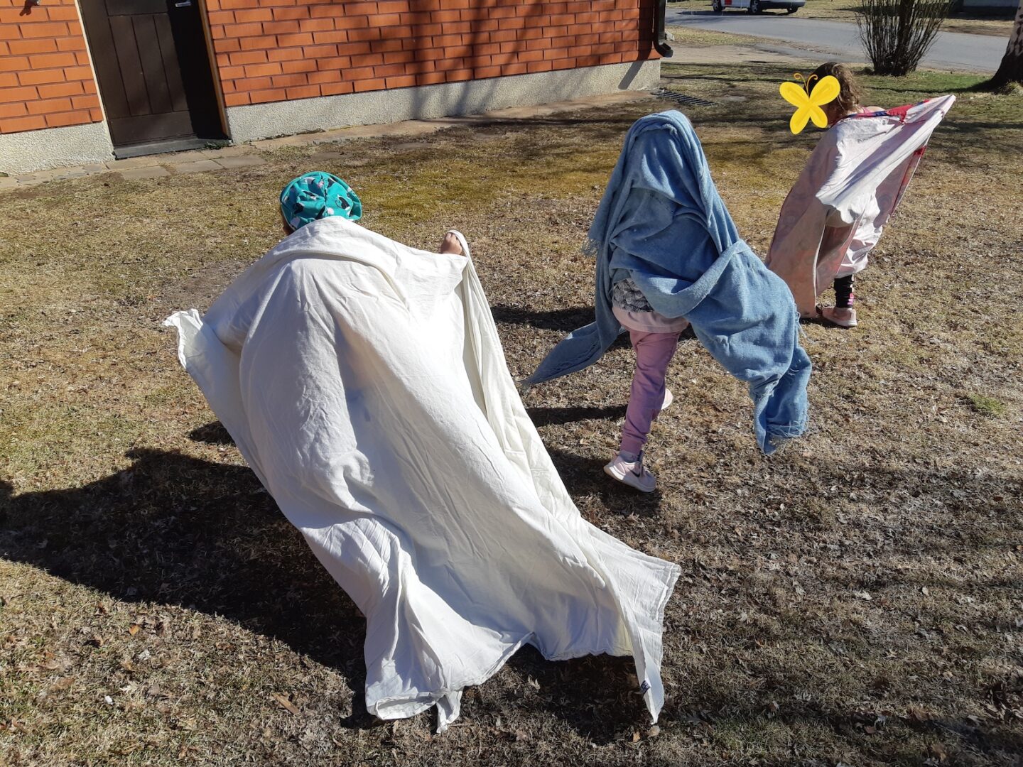 Kolme lasta leikkii ulkona lakanat päällään.