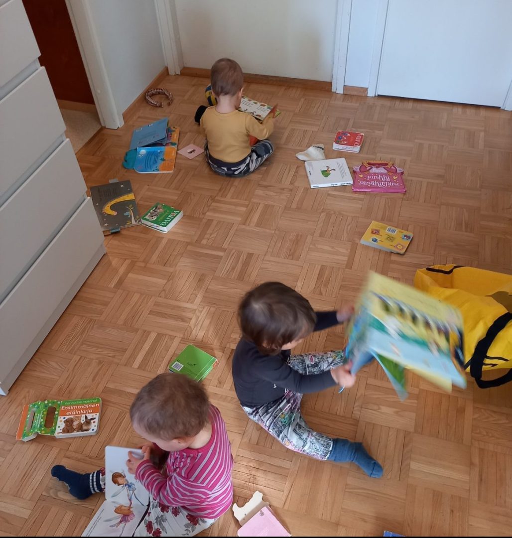 Kolme lasta istuu lattialla kirjojen keskellä ja lukee niitä.