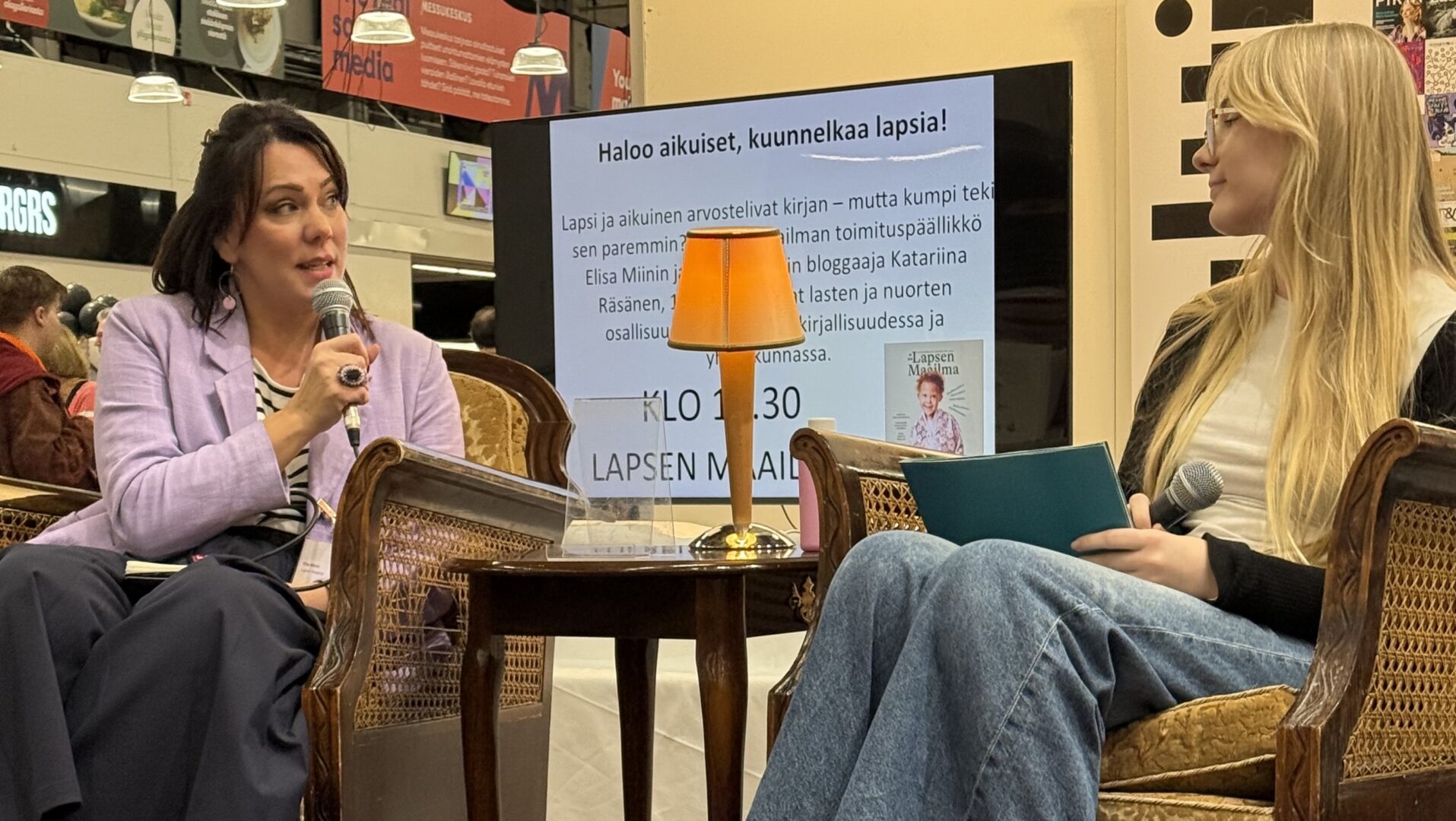 Kaksi naista keskustelee pienen pöydän ääressä Kirjamessuilla.
