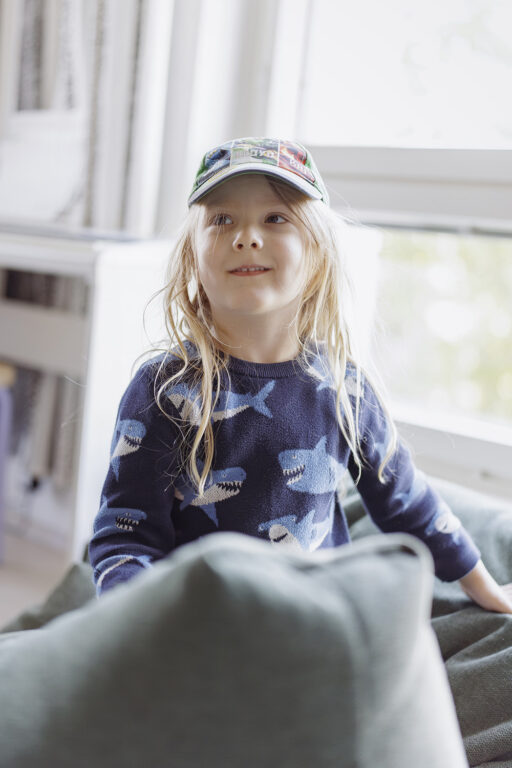 Pieni poika istuu polvillaan sohvalla. Hänellä on päässään lippalakki ja päällä haikuvioinen paita. 