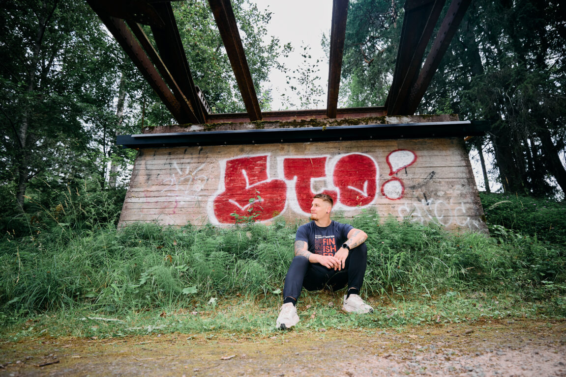 Jouni Kokkonen istuu metsässä vanhan tiiliseinän edessä. Taustalla on graffiti.