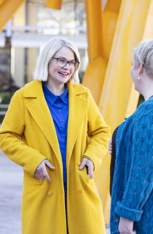 Kati Leskinen seisoo ulkona ja katsoo Annika Saarikkoa nauraen.