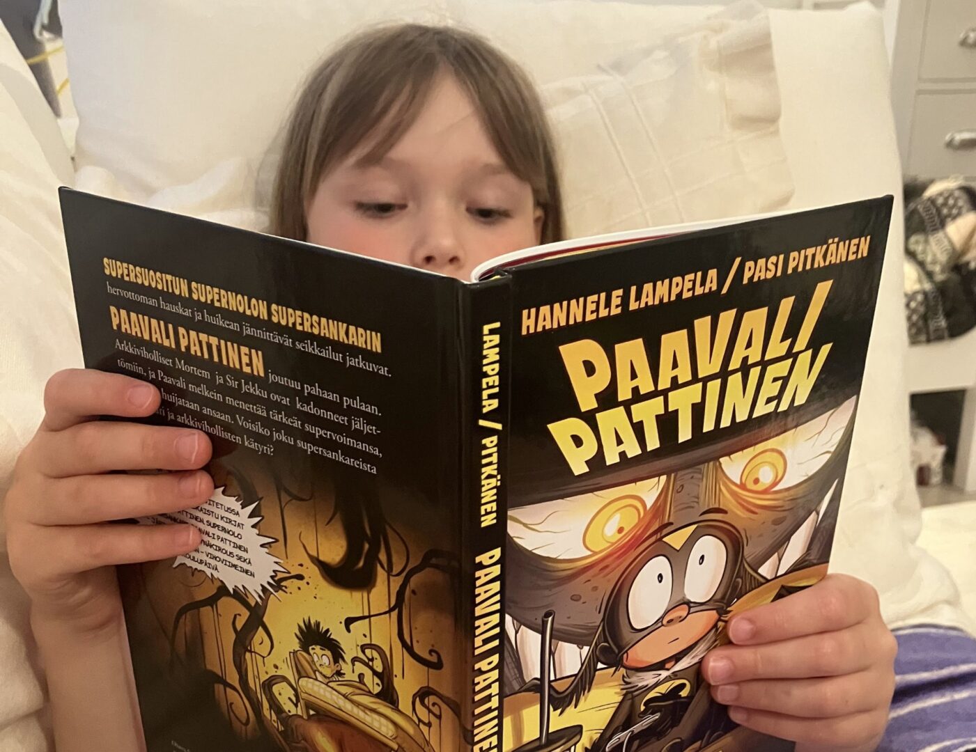 Tyttö istuu nojatuolissa ja pitää kasvojensa edessä kirjaa Paavali Pattinen.
