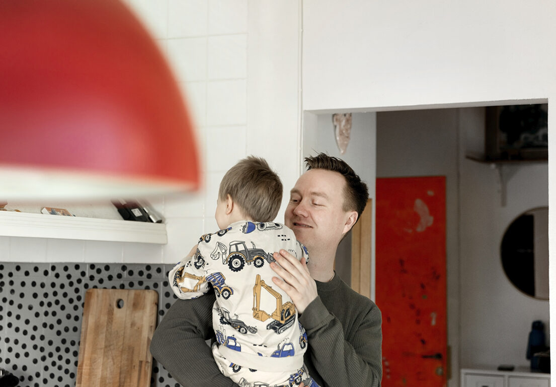 Ville Viholainen pitää sylissään leikki-ikäistä lasta, jonka vaatteissa on työkoneiden kuvia.